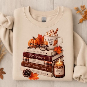 May include: A beige crewneck sweatshirt featuring a graphic print of a stack of books, a mug with whipped cream and text reading 'hello fall', a pumpkin, eyeglasses, a pinecone, and a candle with a label that reads 'sweater weather'.