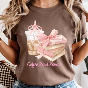以下が含まれることがあります： コーヒーの飲み物、ピンクのリボンで結ばれた本の山、そして「Coffee Read Repeat」のテキストの水彩画グラフィックが描かれた茶色のTシャツ。カジュアルなスタイルのシャツです。