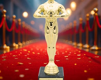 Könnte beinhalten: Eine goldfarbene Trophäenstatue auf einem schwarzen Sockel, die auf einem roten Teppich steht. Die Statue zeigt eine stilisierte Figur, die ein Schwert hält. Der Sockel hat ein goldenes Schild mit deutschem Text. Im Hintergrund ein roter Teppich und Samtseile.