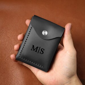 May include: A black leather card holder featuring a flap closure with a silver-toned metal snap button. The front is embossed with the letters M|S in a serif font. The edges are secured with visible black stitching along the side and bottom.