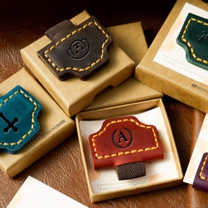 Puede incluir: Marcadores de cuero en varios colores, incluyendo marrón, verde azulado y rojo, con costuras amarillas. Cada marcador se presenta en una pequeña caja de cartón. Algunos presentan diseños en relieve, como un mono o la letra "A".