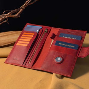 Puede incluir: Portapasaporte de cuero rojo abierto con múltiples ranuras para tarjetas, un bolsillo con cremallera y un cierre a presión. El soporte contiene un pasaporte y un pasaporte británico. Un AirTag de Apple es visible.