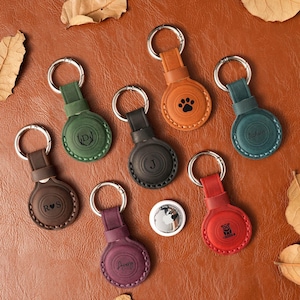 May include: A collection of leather keychains in various colors, including brown, green, orange, and red. Each keychain features a circular design with a silver keyring and personalized engravings, such as initials, names, and a paw print. An Apple AirTag is also visible.