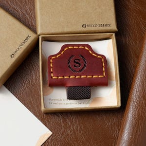 Puede incluir: Un marcapáginas de cuero burdeos con un borde dorado cosido y la letra "S" dentro de una corona de laurel. El marcapáginas está en una caja de cartón con el logotipo "BYGONEMARK". El texto "If gratitude were...I'd send you a garden in full bloom" es visible.