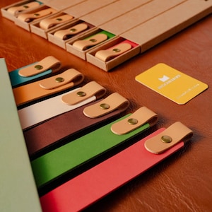 May include: A collection of colorful leather bookmarks with tan leather loops and brass rivets. Some bookmarks are displayed in open cardboard boxes with colored interiors. A yellow card with text is also visible.