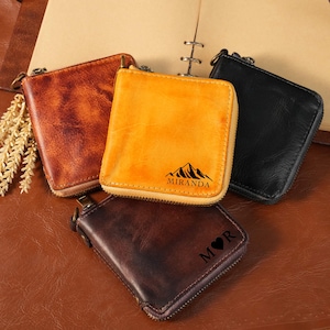 Puede incluir: Cuatro carteras cuadradas con cremallera en tonos marrón claro, amarillo, marrón oscuro y negro. La cartera amarilla presenta un gráfico de montaña con el texto MIRANDA. La cartera marrón oscuro tiene grabadas las letras M, un corazón y R.