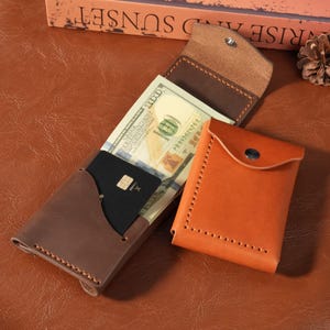May include: A dark brown leather wallet with visible stitching, containing a one hundred dollar bill and a black credit card, positioned next to a smaller, folded tan leather wallet with a silver snap button closure and matching stitching.