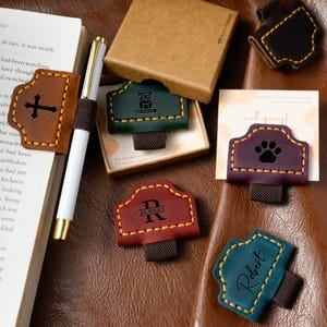 May include: A collection of leather pen holders in various colors, including brown, green, red, purple, and teal. Each holder features unique designs such as a cross, an owl, a paw print, and personalized text. The holders are displayed on a brown leather surface.