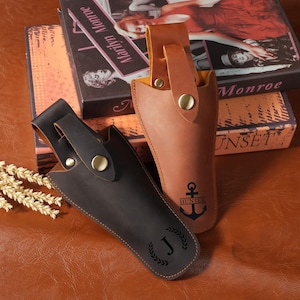 Puede incluir: Dos fundas de cuero con trabillas para cinturón y cierres a presión de color latón. La funda marrón oscuro presenta una letra J dentro de una corona de laurel. La funda marrón claro muestra un gráfico de ancla sobre el texto HUNTER en negro.