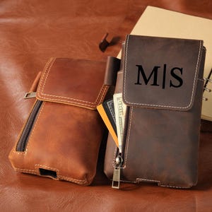Puede incluir: Dos fundas de cuero para cinturón en marrón claro y oscuro. La funda oscura tiene una solapa con las letras M|S grabadas, un bolsillo frontal con cremallera y una ranura abierta que contiene una tarjeta de crédito y un billete de cien dólares.