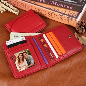 Puede incluir: Cartera de cuero roja abierta, con múltiples ranuras para tarjetas, una ventana de identificación transparente con una foto y un cierre a presión. La cartera contiene tarjetas y efectivo. También se ve una cartera roja más pequeña a juego.