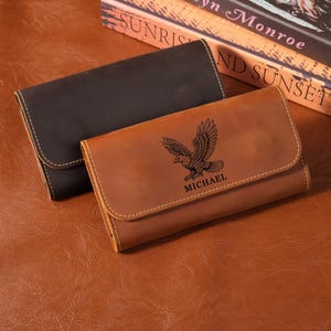 Puede incluir: Dos carteras rectangulares de cuero con cierre de solapa y costuras visibles. Una cartera es de color marrón oscuro y la otra es de color tostado con un gráfico negro grabado de un águila en vuelo sobre el nombre MICHAEL en la solapa.