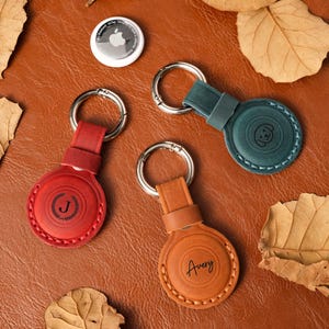 Puede incluir: Tres llaveros circulares de cuero con anillas metálicas, uno rojo con la letra J, uno color canela con el nombre Avery y uno verde azulado con un icono de cara de perro. Un AirTag de Apple blanco se sitúa encima sobre una superficie marrón.