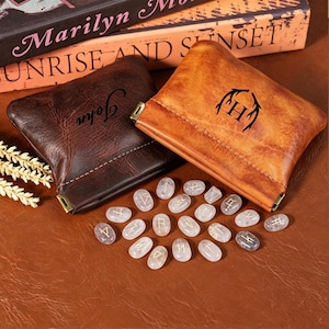 Puede incluir: Dos bolsas de cuero, una marrón con "John" y otra marrón claro con un diseño de cuerno negro y la letra "H". Piedras rúnicas ovaladas y transparentes con símbolos dorados esparcidas. Sobre una superficie marrón.
