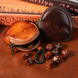 Op de afbeelding: Een bruine leren dobbelsteenzak met een rits, naast een kleinere bruine leren etui met de letter "H" en geweien. Verschillende bruine dobbelstenen met gouden cijfers liggen verspreid rond de tassen. De tas is open.
