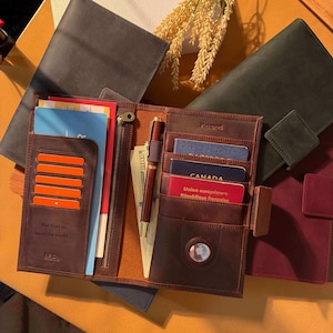 Puede incluir: Cartera de viaje de cuero marrón abierta con múltiples compartimentos para tarjetas, pasaportes y efectivo. Incluye un bolígrafo y un bolsillo con cremallera. La cartera tiene un cierre de botón a presión y el texto "For God so loved the world..." es visible.