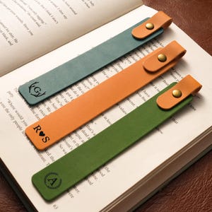 Puede incluir: Tres marcapáginas de cuero en verde azulado, naranja y verde, cada uno con una lengüeta de cuero marrón claro y un broche de latón. Los marcapáginas están personalizados con iniciales y un emblema decorativo, sobre las páginas de un libro abierto.