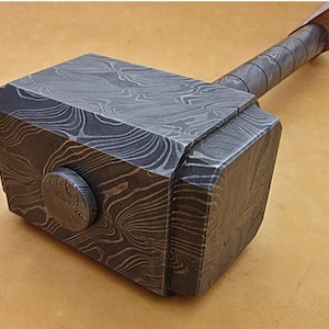 Könnte beinhalten: Ein handgefertigter Metallhammer mit einem gemusterten Design auf dem Kopf und einem umwickelten Griff. Der Hammerkopf ist dunkelgrau mit einem wirbelnden, holzmaserungsartigen Muster. Der Griff ist mit einem dunkelgrauen Material umwickelt.
