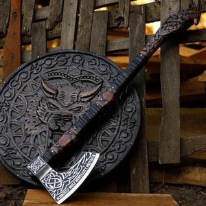 May include: A handcrafted Viking axe and shield set. The axe features a detailed wolf head carving on the handle and a silver blade with engraved text. The shield is round, dark brown, and has a wolf design in the center. The set is leaning against a wooden fence.