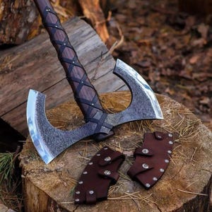 May include: A Viking-style axe with a dark wooden handle wrapped in black leather. The axe head is silver and curved, with a textured finish. Two brown leather sheaths with silver rivets are included. The axe rests on a wooden surface.