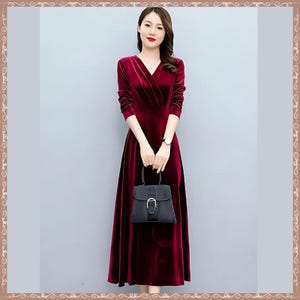 May include: A long, burgundy velvet dress with a V-neck and long sleeves. The dress is paired with a black handbag with a silver buckle. The woman is wearing a silver watch. The dress has a flowing skirt.