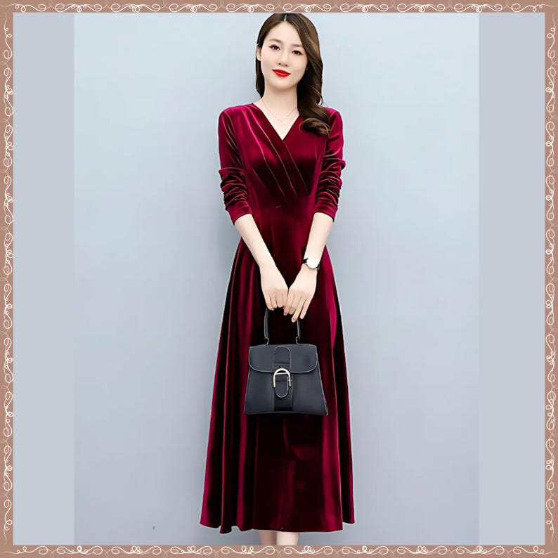 May include: A long, burgundy velvet dress with a V-neck and long sleeves. The dress is paired with a black handbag with a silver buckle. The woman is wearing a silver watch. The dress has a flowing skirt.