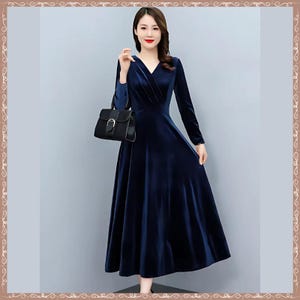 May include: A long, navy blue velvet dress with a V-neck and long sleeves. The dress has a flowing A-line skirt and is paired with a black handbag. The woman is standing in front of a light gray background.