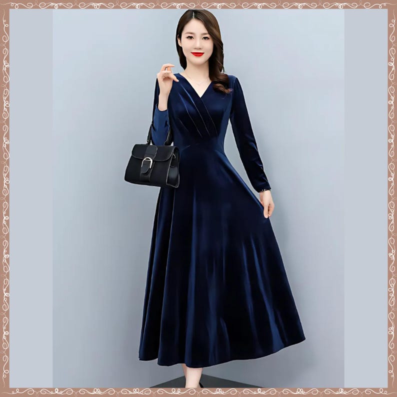 May include: A long, navy blue velvet dress with a V-neck and long sleeves. The dress has a flowing A-line skirt and is paired with a black handbag. The woman is standing in front of a light gray background.