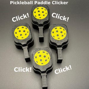 May include: Four black pickleball paddle clickers with yellow ball designs are arranged on a gray surface. The text "Pickleball Paddle Clicker" and the word "Click!" are also visible.