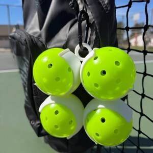 May include: Four neon green pickleballs with white holders are attached to a black carabiner clip. The pickleballs are hanging next to a black sports bag and a black net. The pickleballs have several small holes.