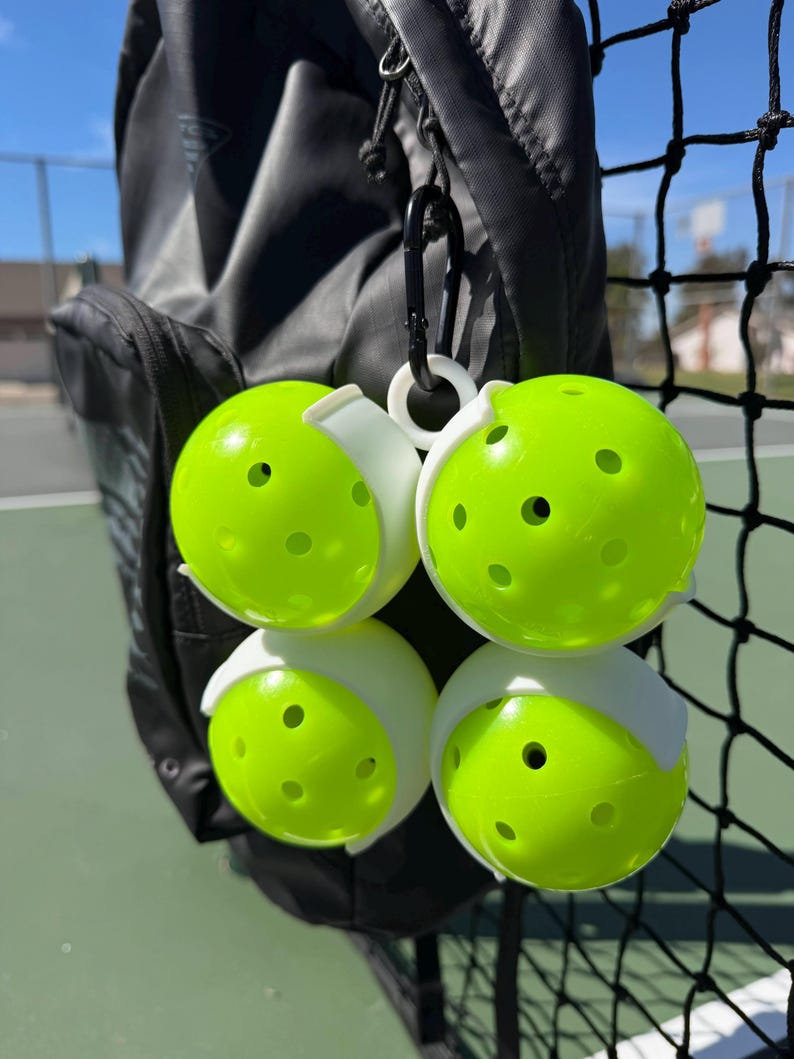 May include: Four neon green pickleballs with white holders are attached to a black carabiner clip. The pickleballs are hanging next to a black sports bag and a black net. The pickleballs have several small holes.