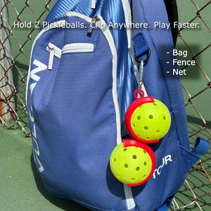 Puede incluir: Una mochila azul con cremalleras blancas y la palabra "VULCAN" impresa. Dos pelotas de pickleball amarillas se sujetan con clips rojos sujetos a la bolsa. El texto "Hold 2 Pickleballs. Clip Anywhere. Play Faster." es visible.
