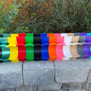 May include: A display of brightly coloured, curved plastic items. The items are arranged in a row and come in a variety of colours, such as black, green, yellow, red, and blue. They are placed on a grey brick surface with green foliage in the background.
