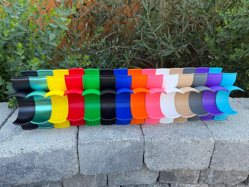 May include: A display of brightly coloured, curved plastic items. The items are arranged in a row and come in a variety of colours, such as black, green, yellow, red, and blue. They are placed on a grey brick surface with green foliage in the background.