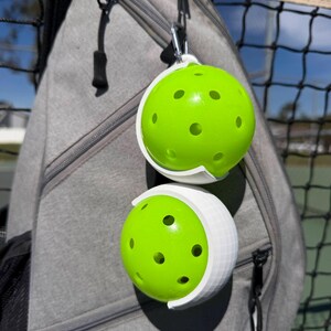 Puede incluir: Dos pelotas de pickleball de color verde brillante con m&uacute;ltiples agujeros se sujetan en soportes blancos sujetos a una mochila gris. Las pelotas de pickleball est&aacute;n sujetas a la mochila con un mosquet&oacute;n plateado. Una red negra es visible en el fondo.