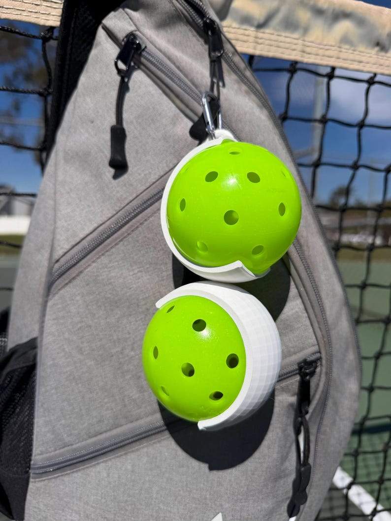 Puede incluir: Dos pelotas de pickleball de color verde brillante con m&uacute;ltiples agujeros se sujetan en soportes blancos sujetos a una mochila gris. Las pelotas de pickleball est&aacute;n sujetas a la mochila con un mosquet&oacute;n plateado. Una red negra es visible en el fondo.