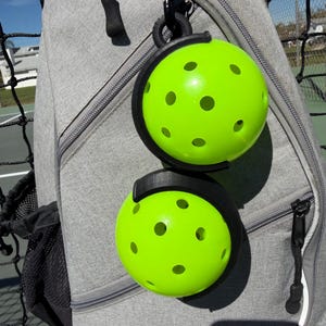 Puede incluir: Dos pelotas de pickleball verde brillante con m&uacute;ltiples agujeros se sujetan en un soporte negro sujeto a una mochila gris. Las pelotas son redondas y tienen un color ne&oacute;n vibrante. La mochila tiene una cremallera y un clip negro.