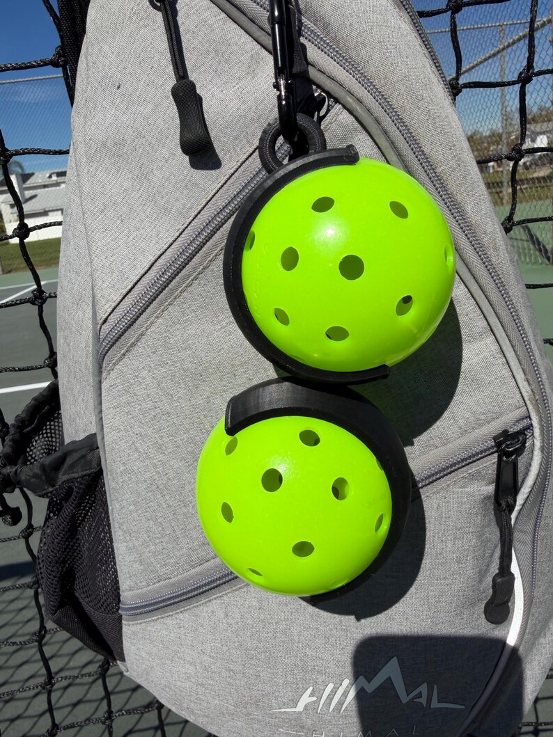 Puede incluir: Dos pelotas de pickleball verde brillante con m&uacute;ltiples agujeros se sujetan en un soporte negro sujeto a una mochila gris. Las pelotas son redondas y tienen un color ne&oacute;n vibrante. La mochila tiene una cremallera y un clip negro.