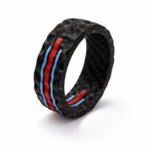 May include: Black ring with a textured surface, featuring three vertical stripes in red and blue. The ring is a band style, with a modern design. The color scheme is reminiscent of racing stripes.
