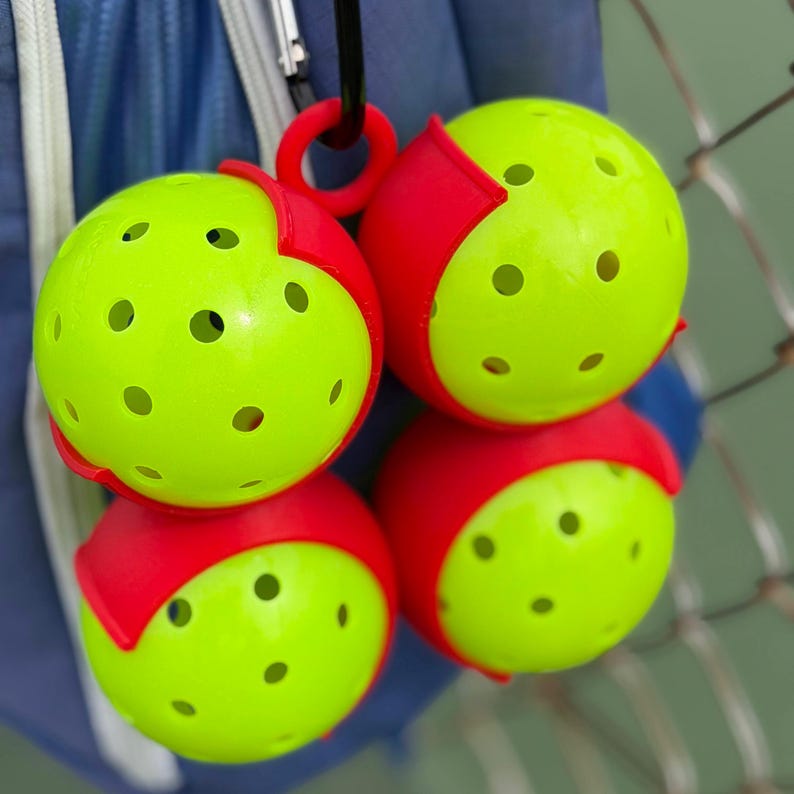 May include: Four pickleballs, each with a bright yellow-green perforated ball and a red curved section. The pickleballs are attached to a black ring and clip, suggesting they are part of a set or accessory for the sport.