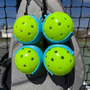 May include: Four neon green pickleballs with multiple holes, each held in a light blue holder. The holders are attached to a grey bag with black carabiners. The background includes a black net and a glimpse of a sunny outdoor setting.