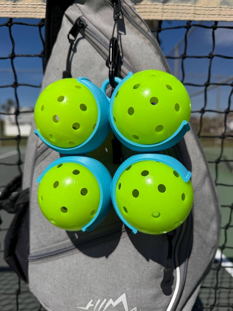 May include: Four neon green pickleballs with multiple holes, each held in a light blue holder. The holders are attached to a grey bag with black carabiners. The background includes a black net and a glimpse of a sunny outdoor setting.
