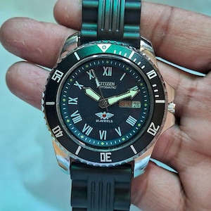 May include: A Citizen automatic watch with a black dial, luminous hands, and a date display. The watch has a black bezel with white markings and a black rubber strap. The watch is held in a hand.