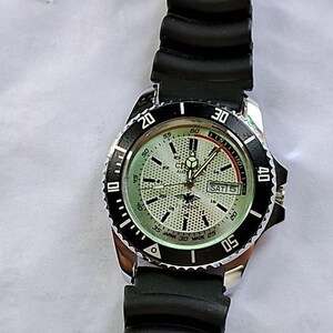 May include: A silver-toned wristwatch with a black bezel and a black rubber strap. The watch face is light green with black and white markings, including the day of the week. The watch has a rotating bezel with white numerals.