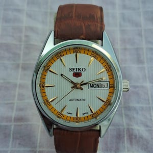 May include: A silver Seiko automatic wristwatch with a white face, gold hands, and a brown leather band. The watch face has gold and orange accents, with the day and date displayed. The watch reads "MON 15".