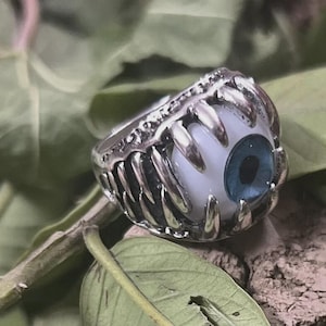 May include: A silver-colored ring featuring a detailed design of a monster's mouth with sharp teeth. The ring holds a large, realistic-looking eye with a blue iris and a white sclera. The ring is set against a natural background.