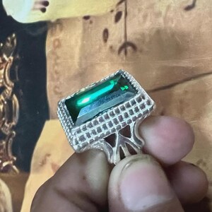 May include: A silver ring with a large, rectangular emerald-cut green gemstone. The ring has a detailed, textured setting around the stone and a decorative band. The ring is held up by a person's fingers.