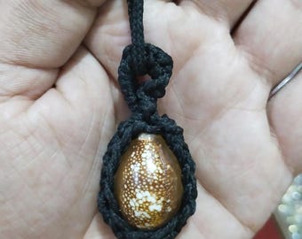 Puede incluir: Un collar artesanal con un colgante de concha ovalada. La concha tiene un patrón moteado marrón y blanco. Está engarzada en un tejido de macramé negro y suspendida de un cordón negro. El colgante mide unos 3 cm de largo.