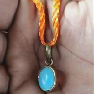Op de afbeelding: Een gouden hanger met een ovale, lichtblauwe steen hangt aan een gevlochten oranje en gele koord. De hanger heeft een eenvoudig ontwerp en de steen heeft een levendige kleur. Het koord is dik en lijkt van een geweven materiaal te zijn gemaakt.