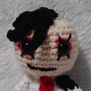 Puede incluir: Muñeca de ganchillo hecha a mano con cabeza color crema, pelo negro y ojos con borde rojo. La muñeca viste camisa blanca, pantalón negro y corbata roja. Los brazos y las piernas de la muñeca también son de color crema.